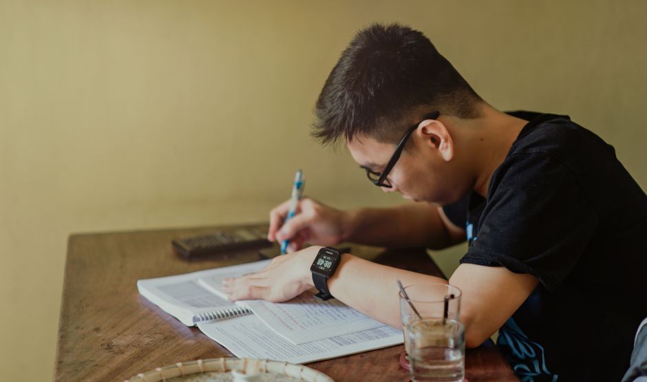 A student studying at a desk with headphones on, looking at a laptop showing a language learning application, surrounded by books and sticky notes.