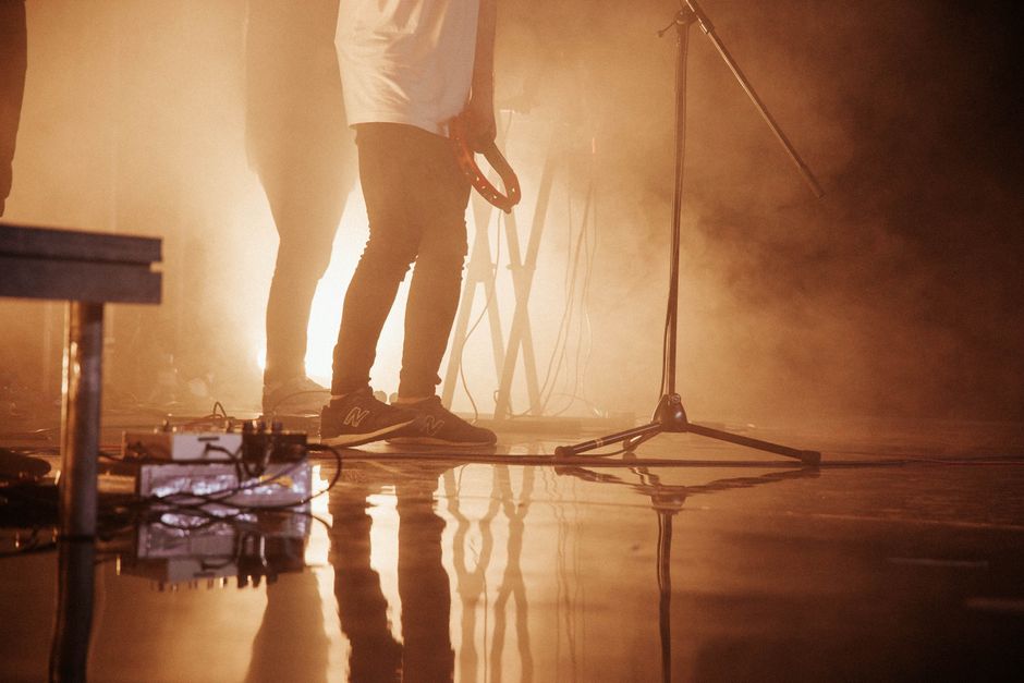 The members of the band What Language Is performing live on a dimly lit stage surrounded by vintage analog synthesizers and acoustic instruments.